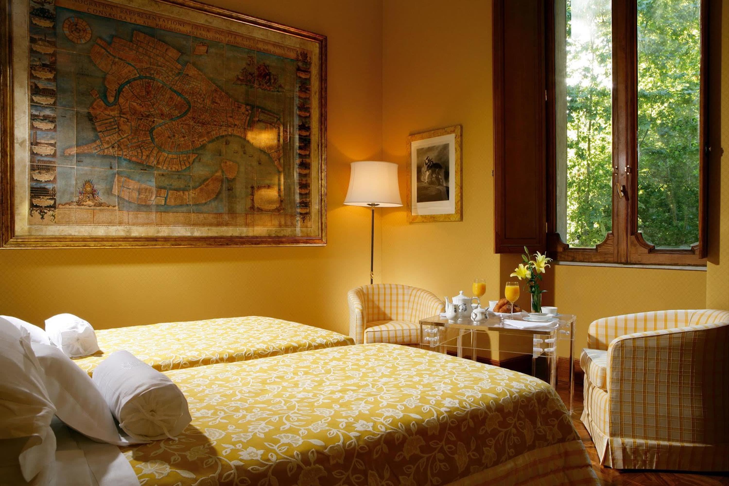 Une chambre d’hôtel avec deux lits, une carte accrochée au mur, une lampe, un tableau encadré et une fenêtre donnant sur des arbres.