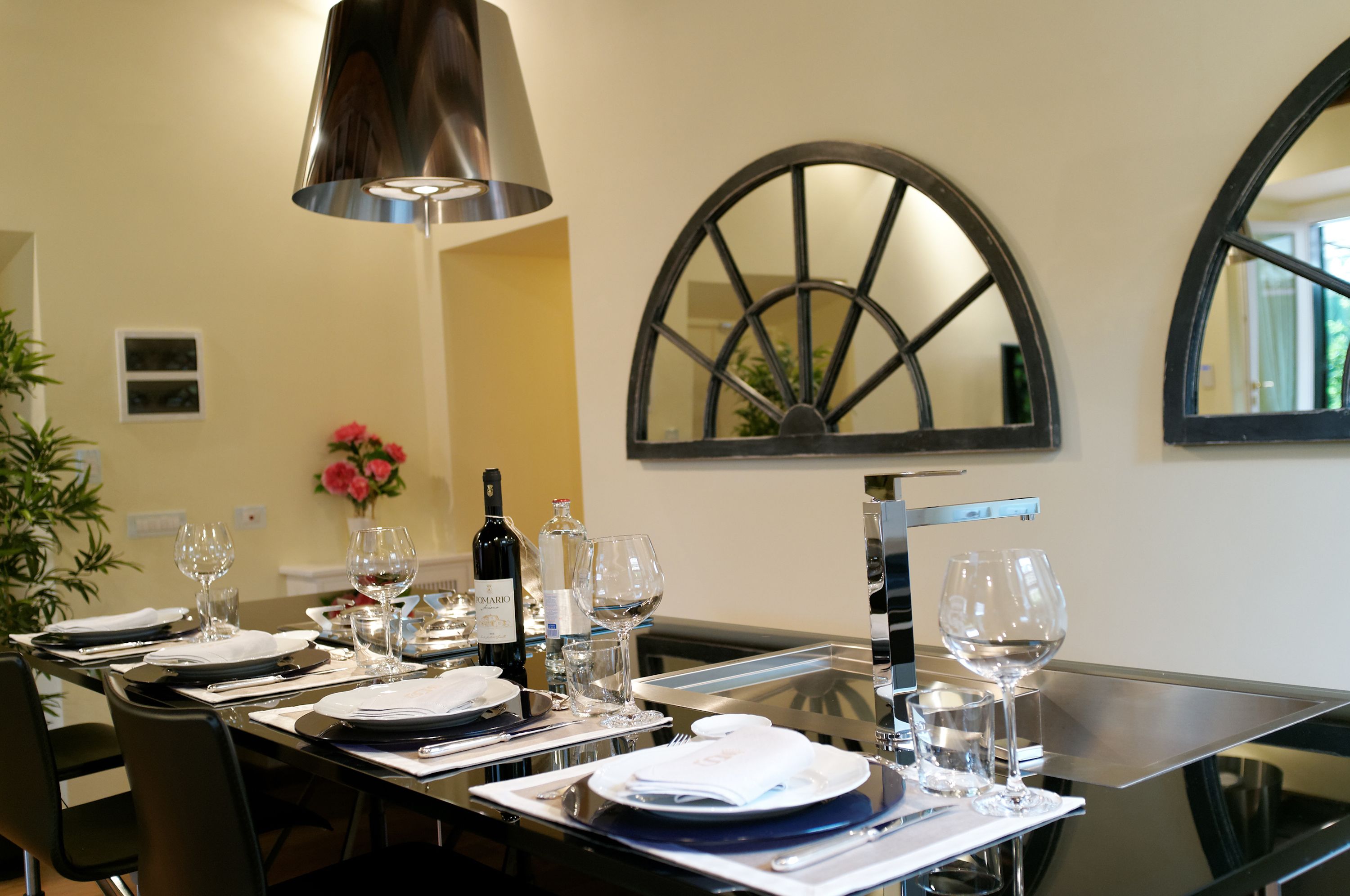 Una mesa de comedor dispuesta con platos, vasos y una botella de vino, con una lámpara moderna y un espejo grande en la pared.