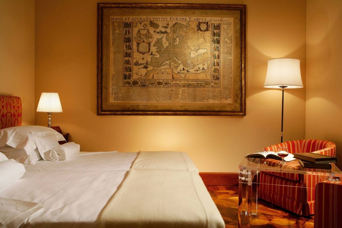 Camera d'albergo con una grande mappa appesa al muro, un letto con lenzuola bianche, un divano a righe rosse e un tavolino trasparente con un libro e degli occhiali.
