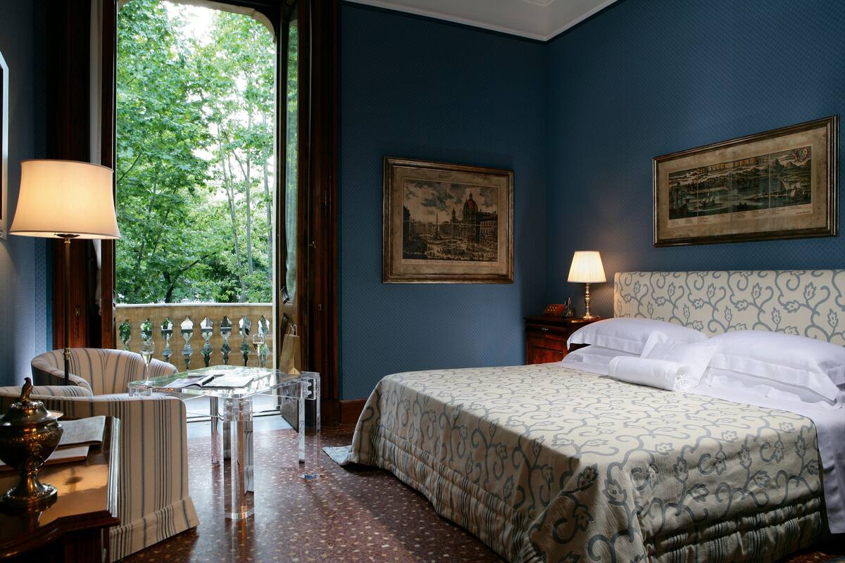 Camera d'albergo con un letto, una sedia, un tavolo e una finestra vista alberi.