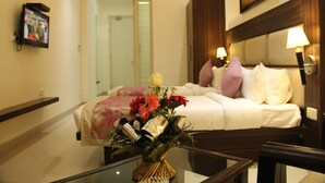 Quarto de hotel com cama, abajures, TV fixada na parede e um vaso com flores sobre a mesa.