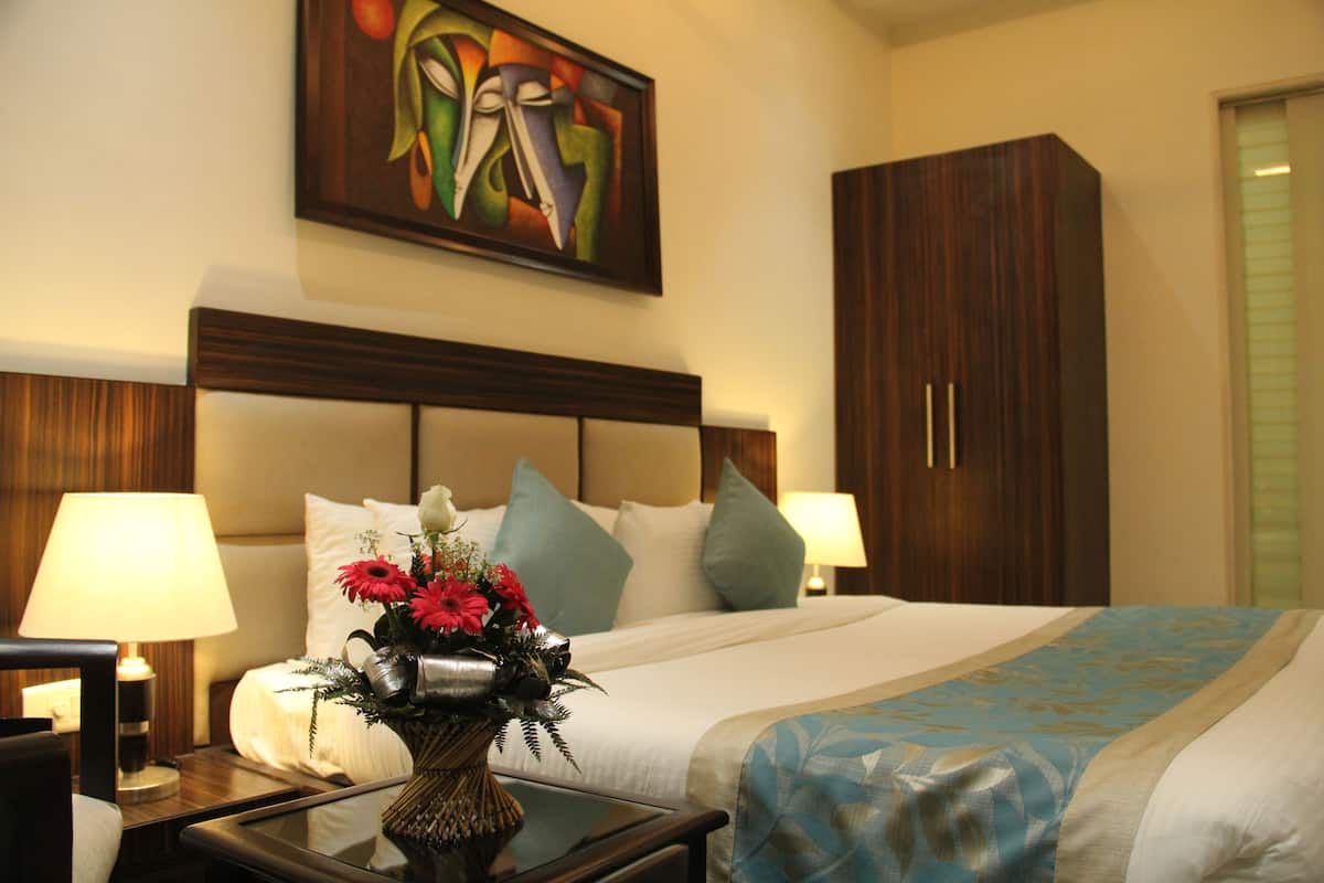 Une chambre d’hôtel avec un grand lit, deux tables de chevet avec des lampes, une table de nuit, un vase décoratif avec des fleurs, un tableau au mur et une armoire.