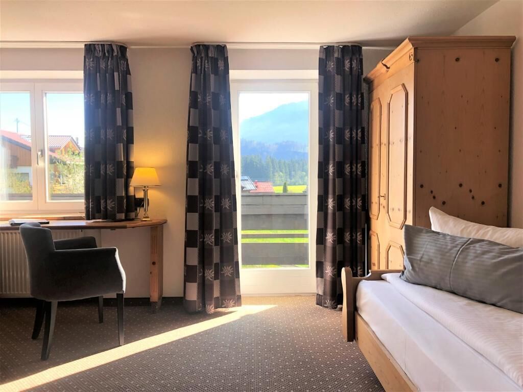 A room with a bed, a desk, and a chair. There is a window with curtains and a view of the outside.
