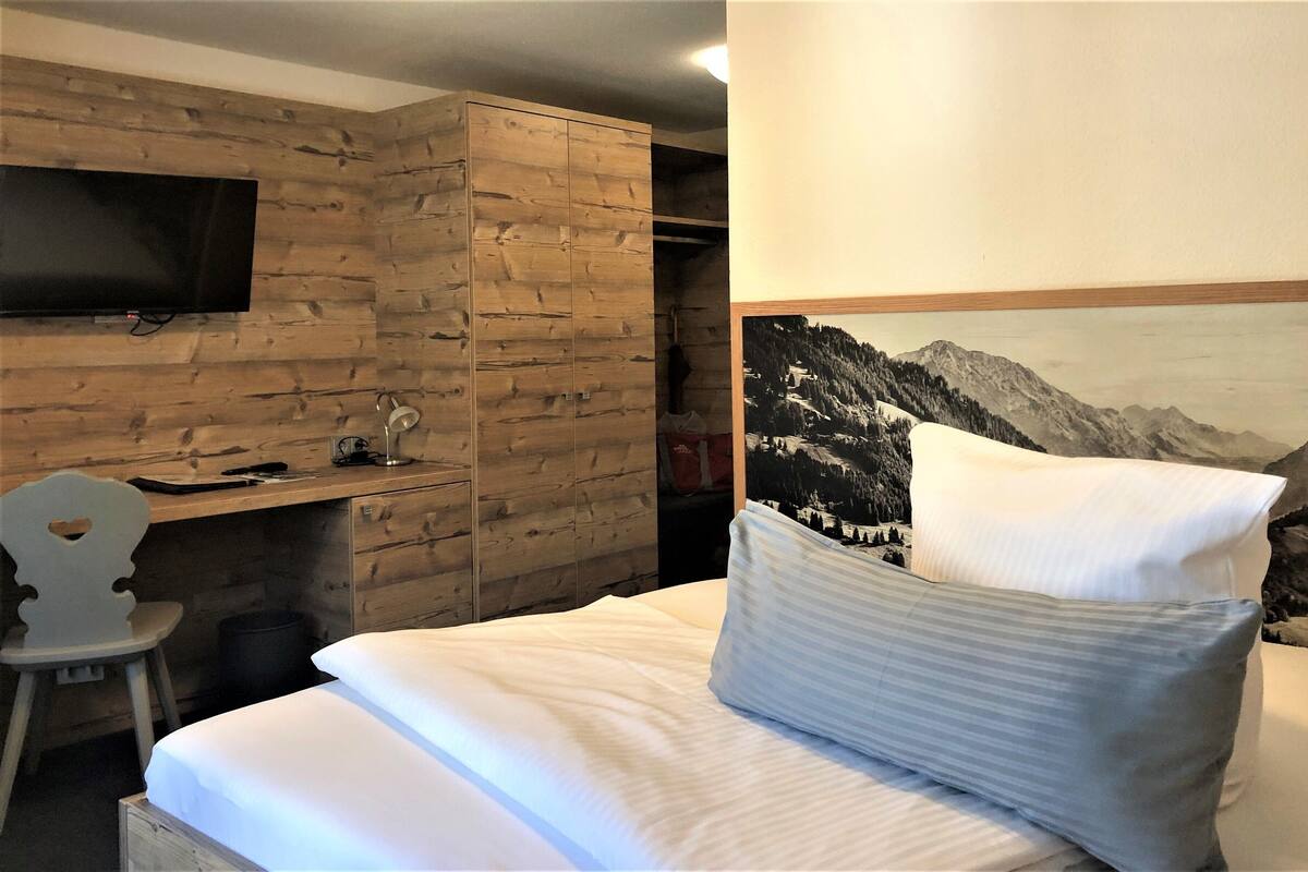 Une chambre d’hôtel avec un mur en bois, un téléviseur fixé au mur, un bureau avec une lampe et un lit agrémenté d’une affiche représentant un paysage de montagne.