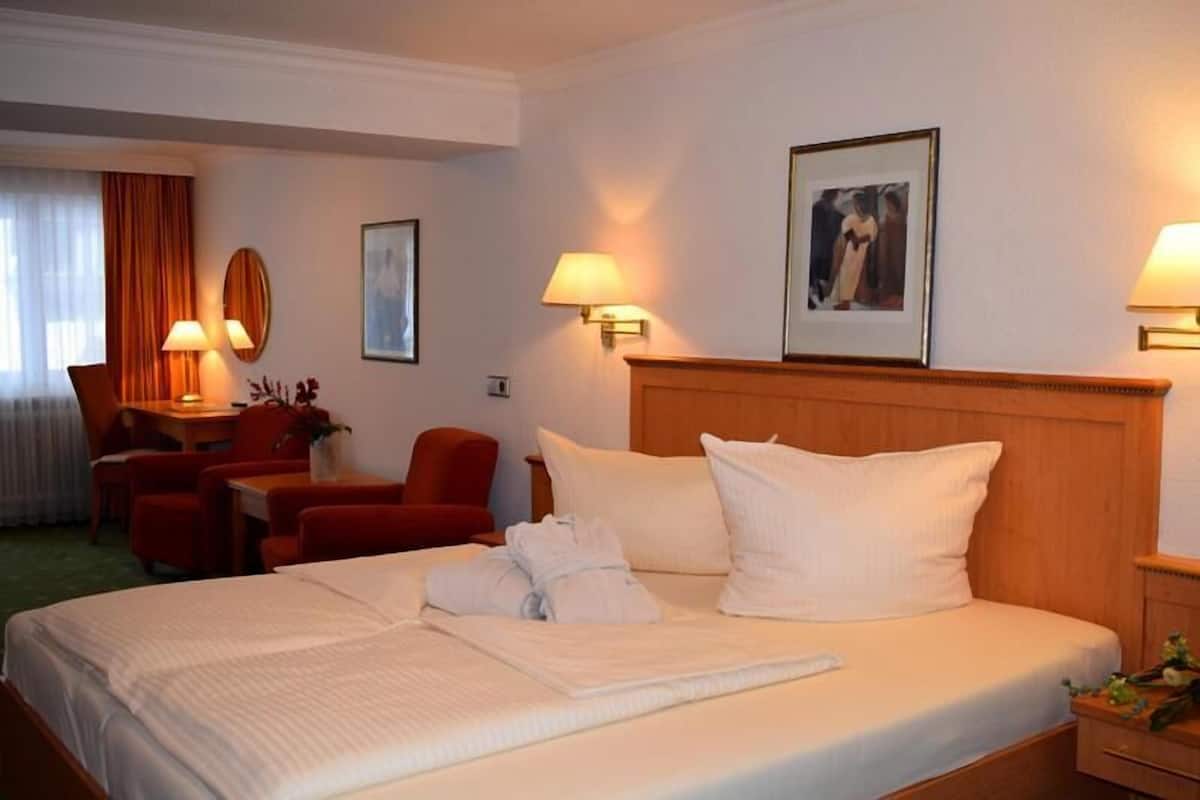 Une chambre d’hôtel avec un grand lit, deux lampes de chevet, une tête de lit en bois, un bureau avec une chaise et une petite table sur laquelle est posé un vase de fleurs.
