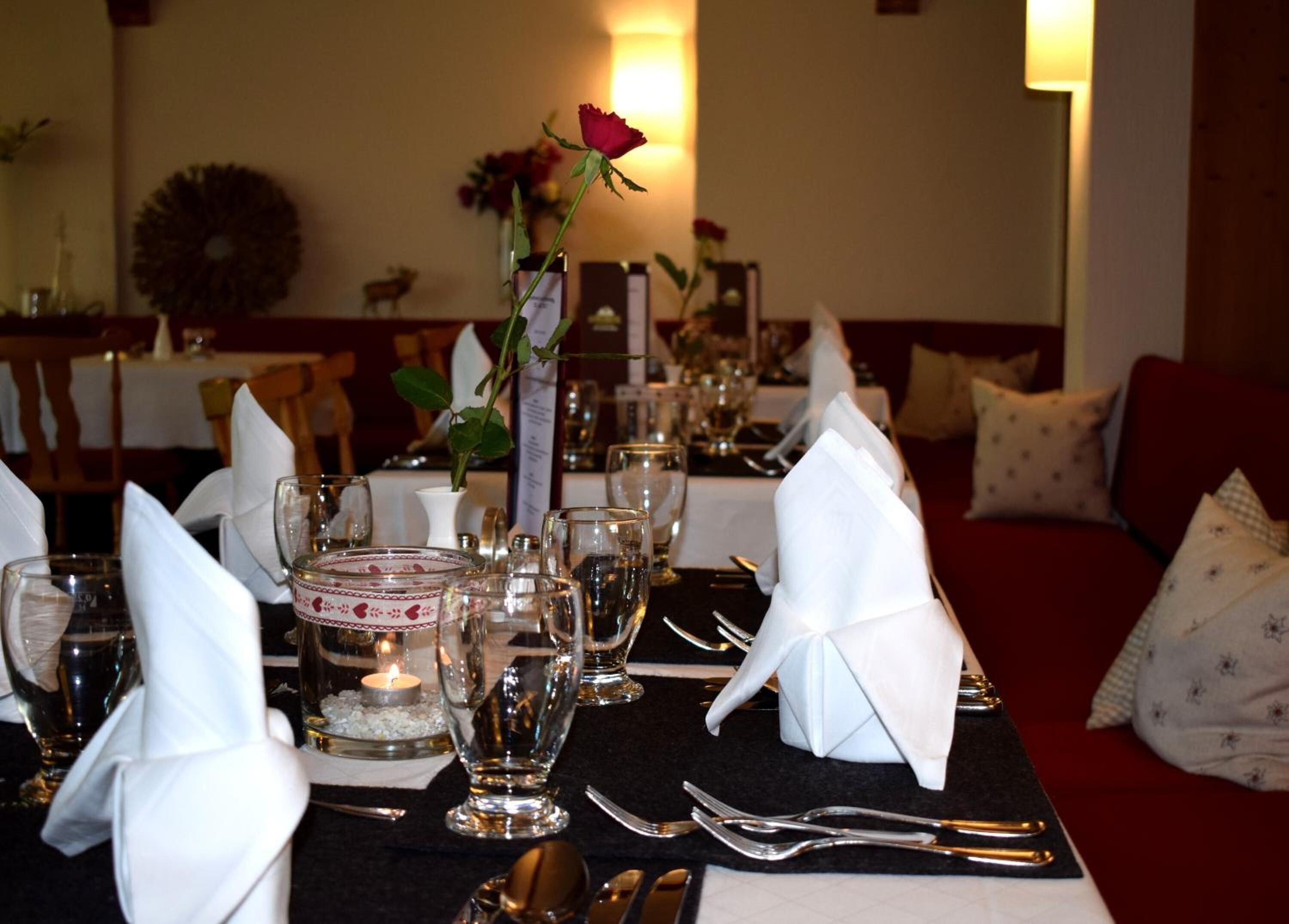 Une table à manger dressée avec des serviettes blanches, des couverts en argent et de la verrerie, agrémentée d’une bougie et d’un bouquet de roses en guise de centre de table.
