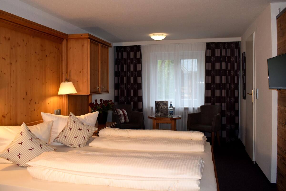 Une chambre d’hôtel avec un grand lit, deux lampes de chevet, une petite table sur laquelle est posé un vase de fleurs, une chaise et un téléviseur fixé au mur.