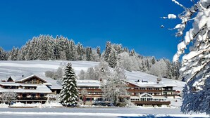 A snow-covered lodge with multiple buildings, a large evergreen tree, and a clear blue sky. - Sonnenbichl Hotel am Rotfischbach (Fischen im Allgaeu)