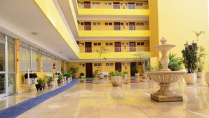 A multi-story building with a yellow facade, a central courtyard with a fountain, potted plants, and a blue carpet leading to the entrance. - Hotel Elegance Palmeras Playa (Puerto de la Cruz)