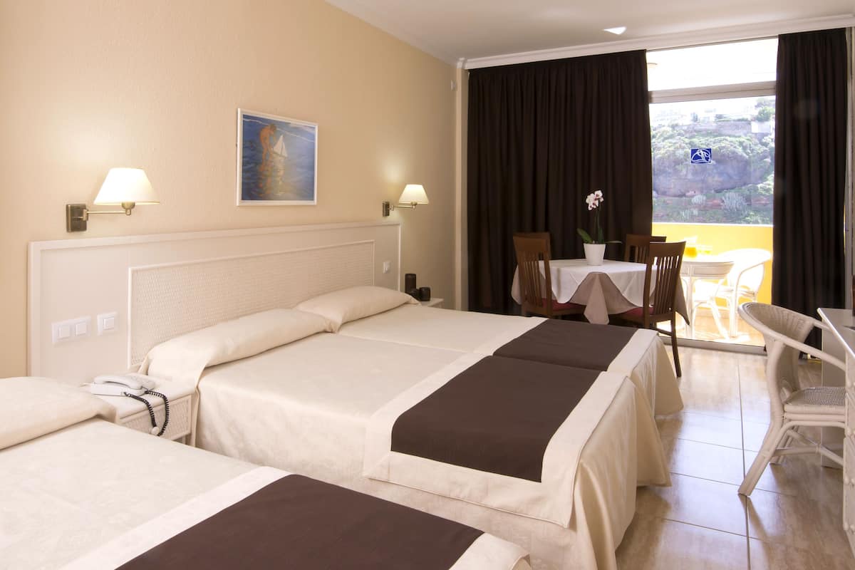 A hotel room with two beds, a desk, a chair, a table, a painting, and a view of the outdoors.