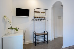 Ein Zimmer mit weißem Konsolentisch, einem schwarzen Metallgestell mit mehreren Kleiderbügeln, einer Topfpflanze und einem an der Wand befestigten Flachbildfernseher.