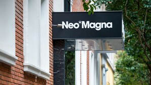 Vista de la calle con un cartel de 'Neo Magna' y árboles podados en macetas a lo largo de la acera.