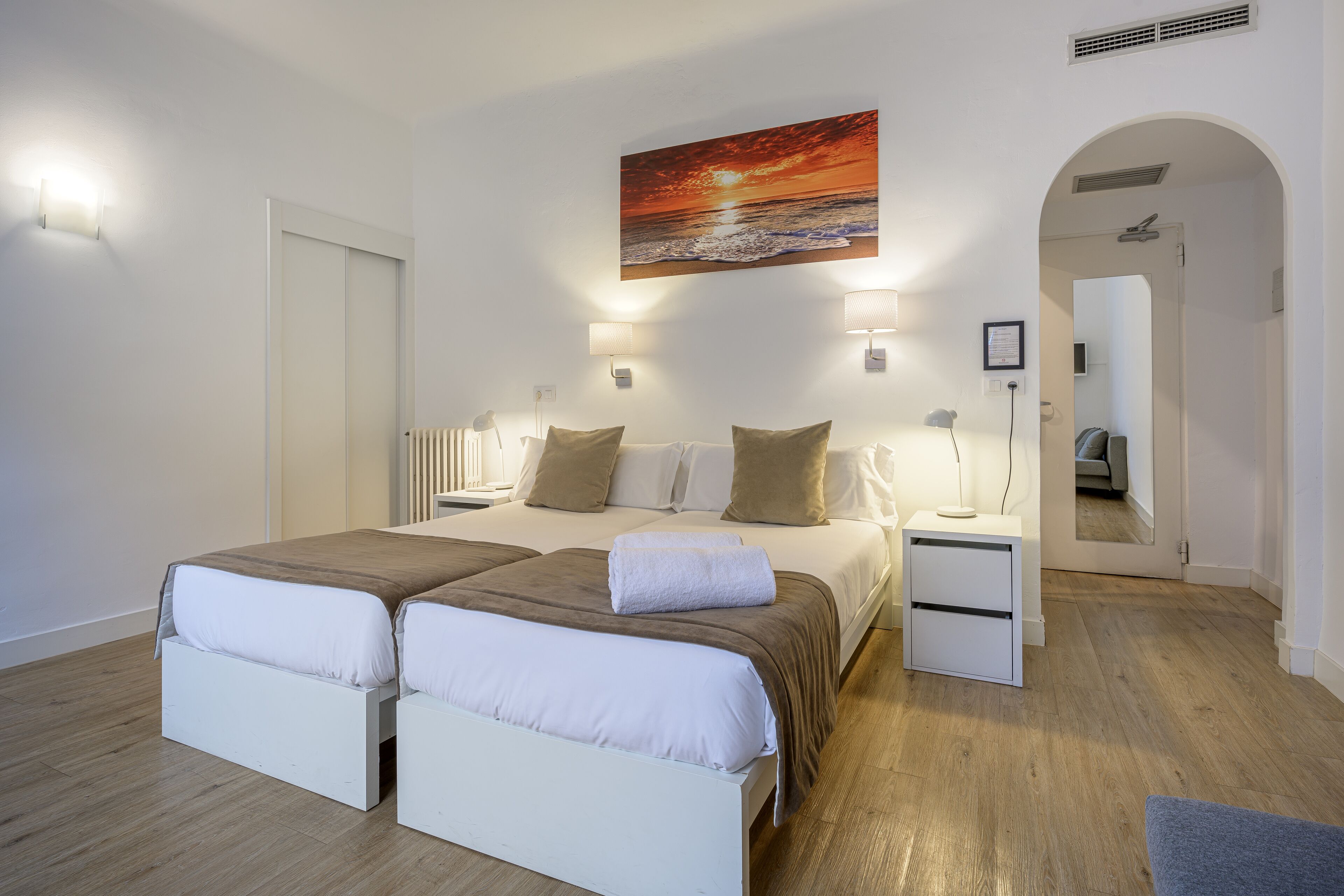 Una cama doble con dos almohadas, una mesita de noche, un bidet y un cuadro colgado en la pared, con una puerta que da a otra habitación.