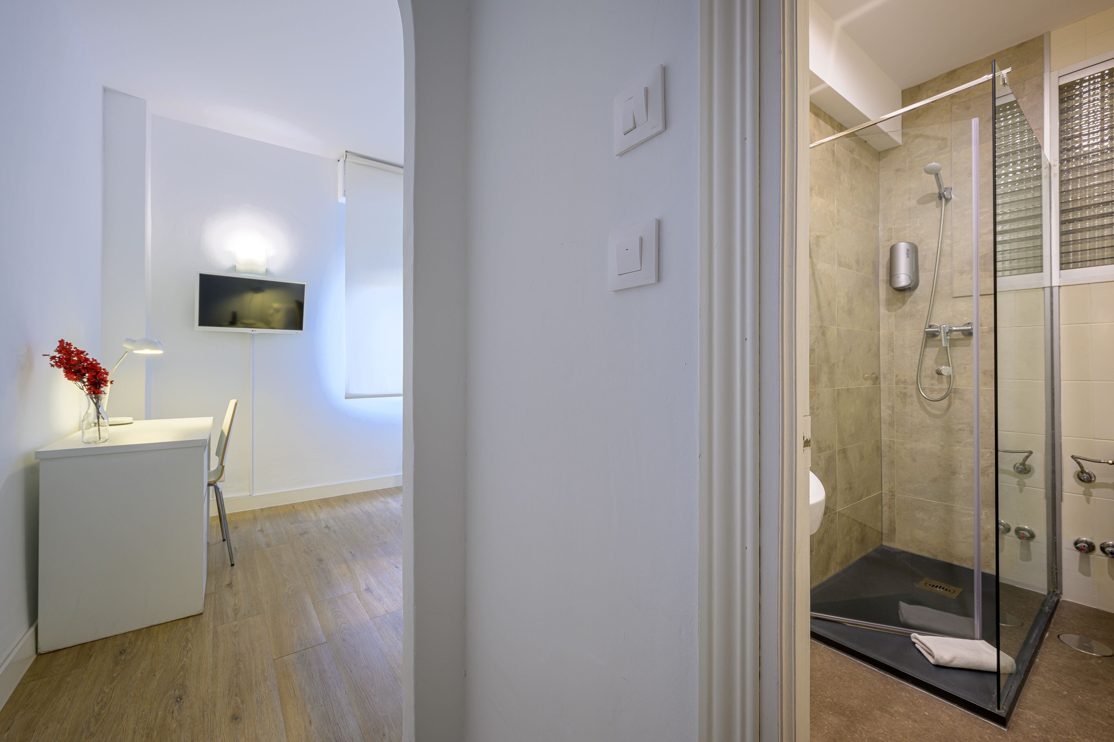 Un baño moderno con cabina de ducha de vidrio, televisor montado y escritorio con un florero.