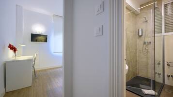 Un baño moderno con cabina de ducha de vidrio, televisor montado y escritorio con un florero.