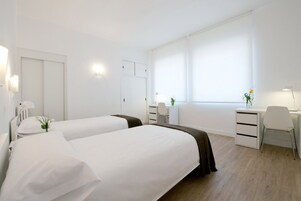 Ein Hotelzimmer mit zwei Betten, einem Schreibtisch, einem Stuhl, einer Kommode und einem Fenster mit Vorhängen.