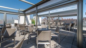 Un área de café al aire libre con sillas y mesas de mimbre, cielo despejado y vistas de edificios al fondo.