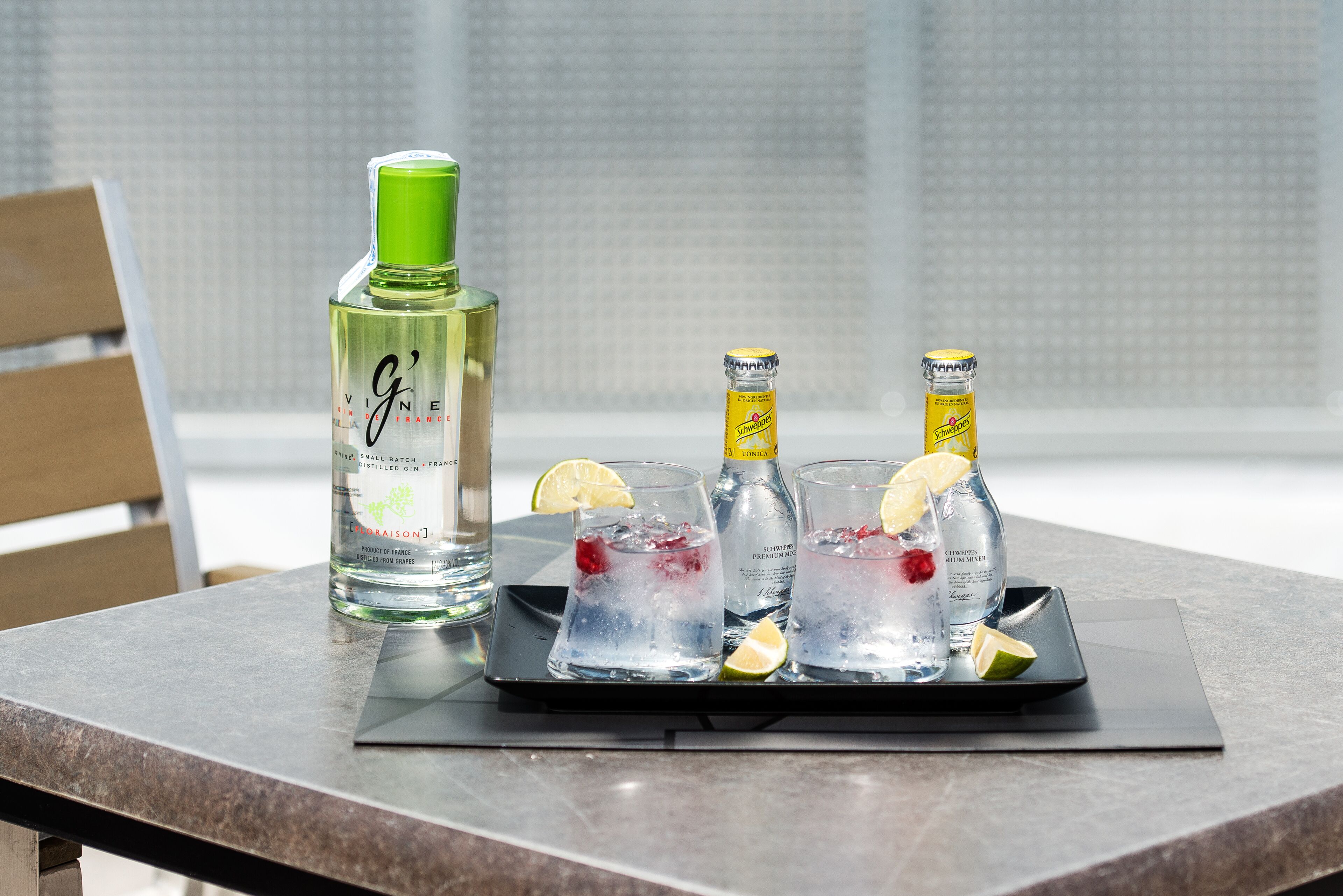 Une assiette sur laquelle se trouvent trois verres remplis d’une boisson, une bouteille de gin et une bouteille d’eau tonique, le tout posé sur une table.