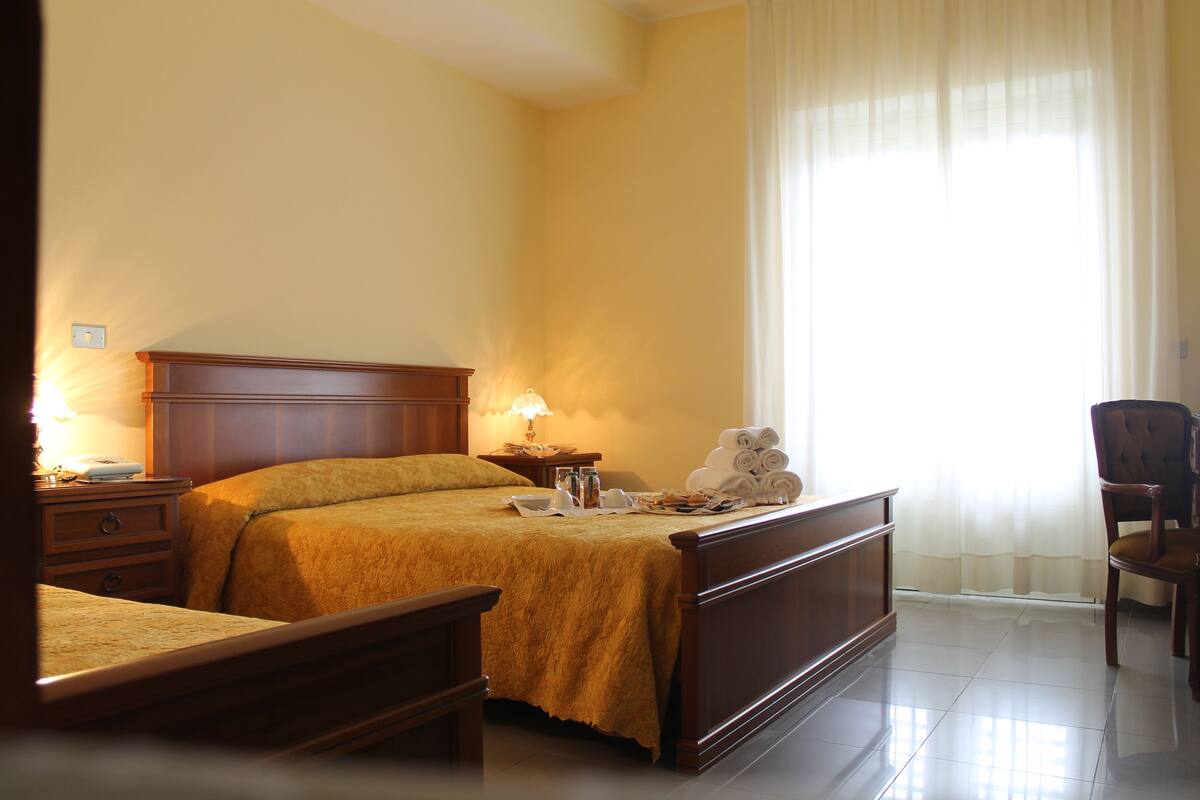 Une chambre d’hôtel avec un lit en bois, une table de chevet avec une lampe, une chaise et une fenêtre avec des rideaux légers.