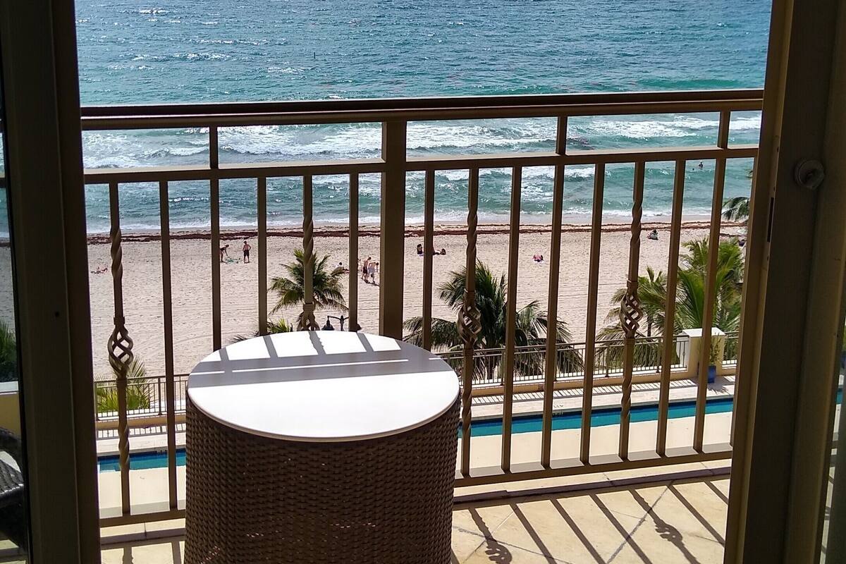 A balcony with a wicker table, a pool, and a beach view.