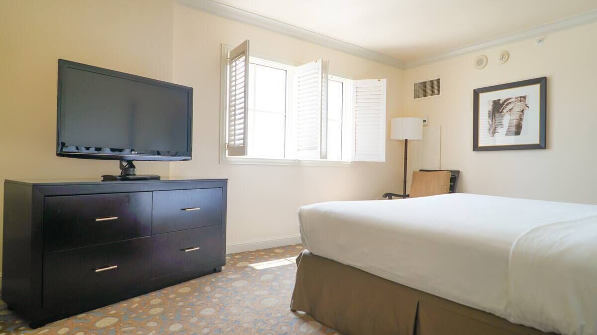 A hotel room with a bed, a TV on a dresser, a window with shutters, and a framed picture on the wall.