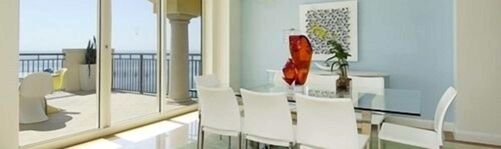 Comedor moderno con mesa de vidrio y sillas blancas, con vista a un balcón y al mar.