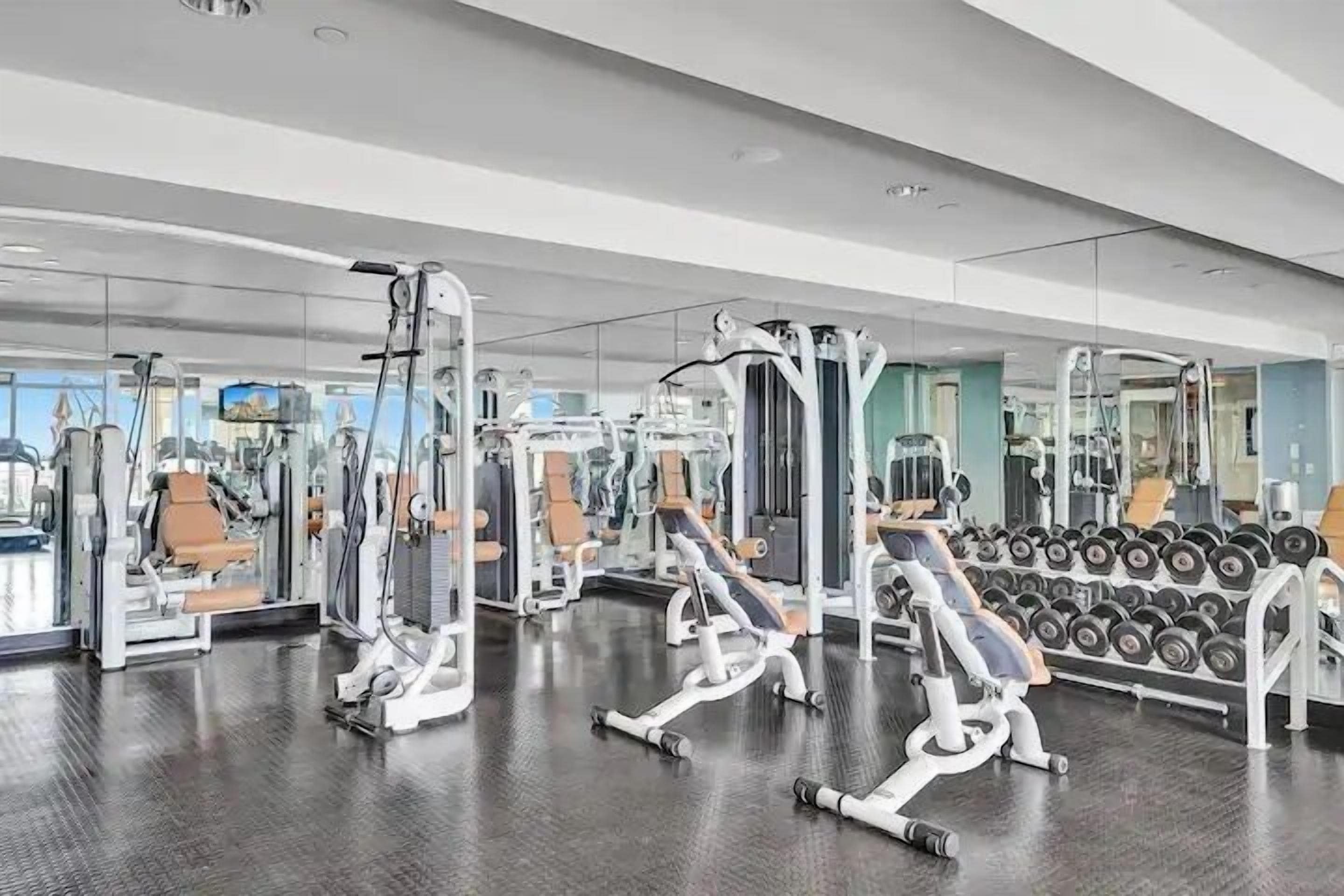 Un gimnasio con diversas máquinas de ejercicio, espejos y un estante con mancuernas.