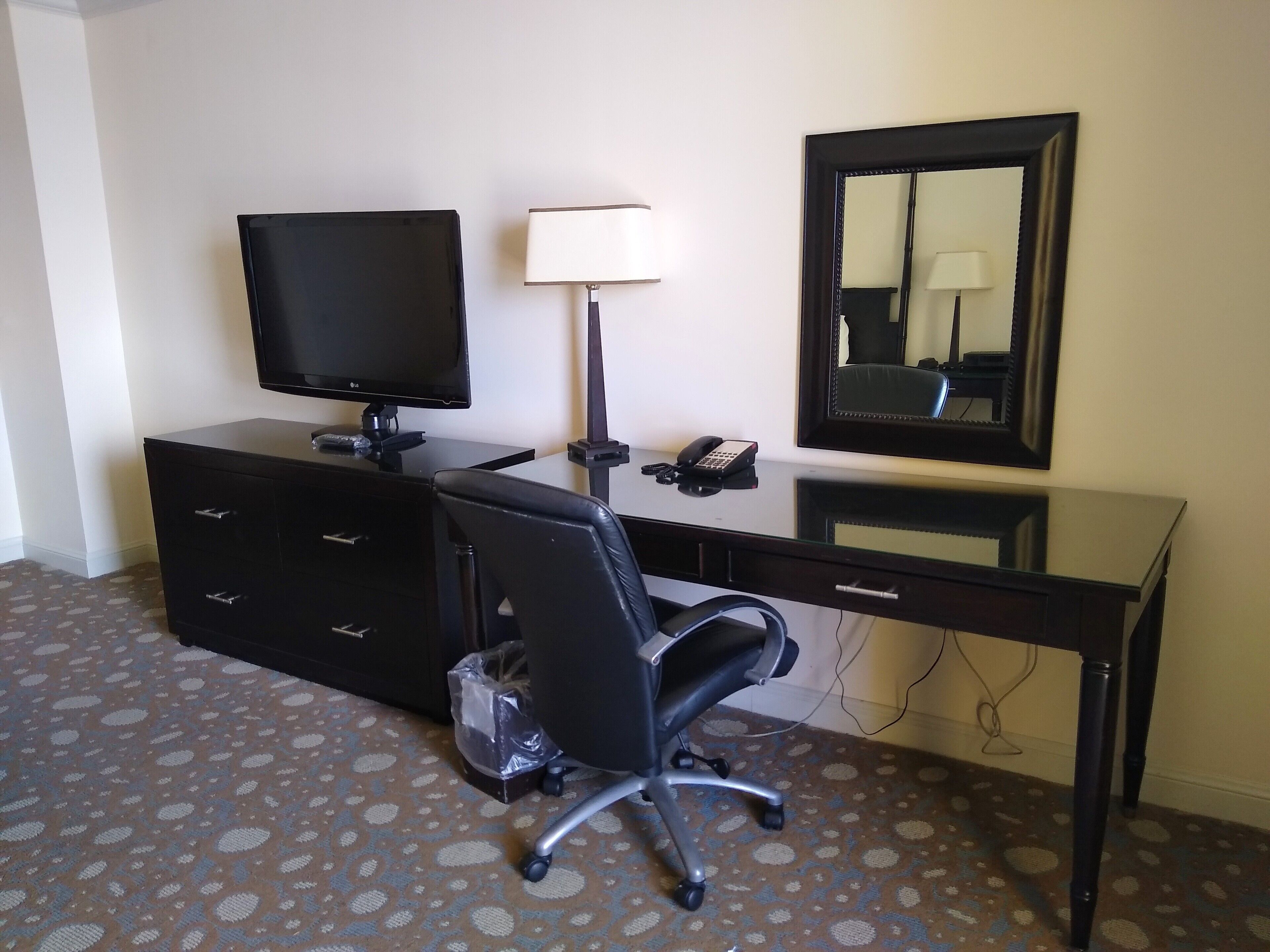 Una habitación de hotel con escritorio, silla, televisor, lámpara, teléfono y espejo.