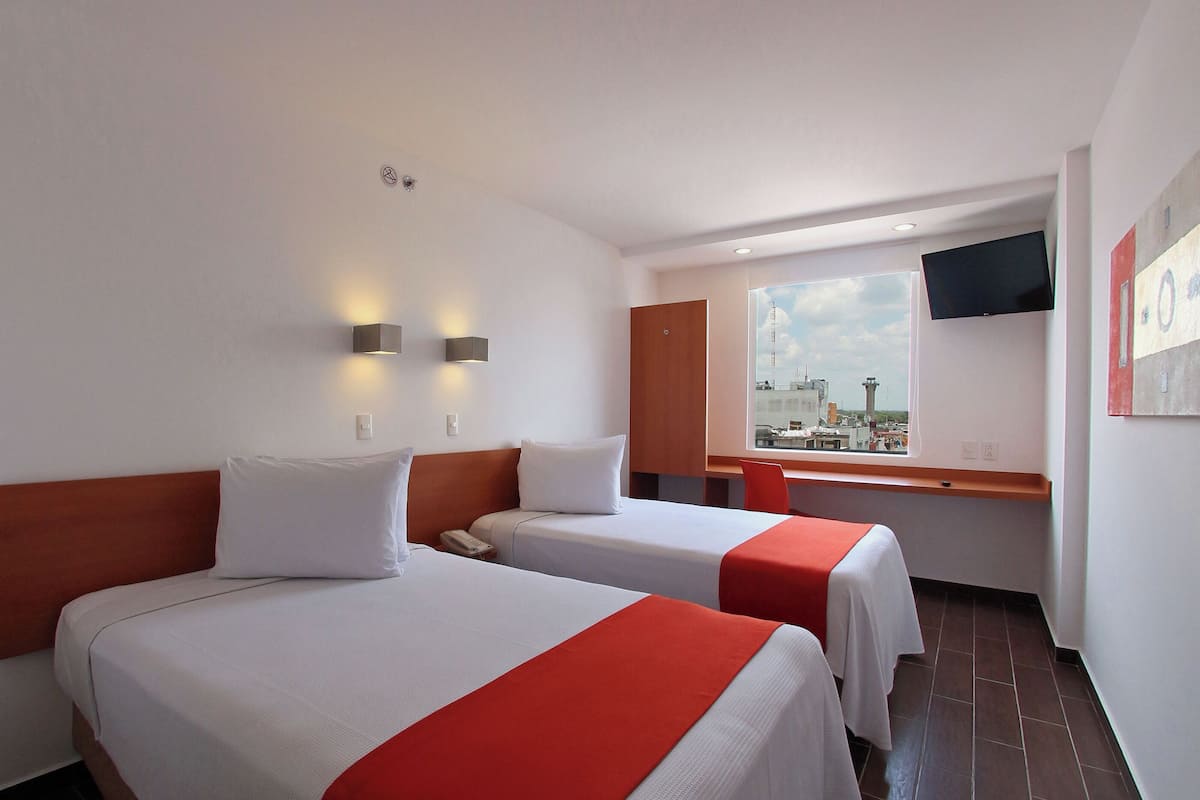 A hotel room with two beds, a TV mounted on the wall, and a window with a city view.