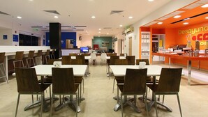 A modern cafeteria with multiple tables and chairs, a counter with food items, and a bright orange wall with signage. - One Villahermosa Centro (Villahermosa)