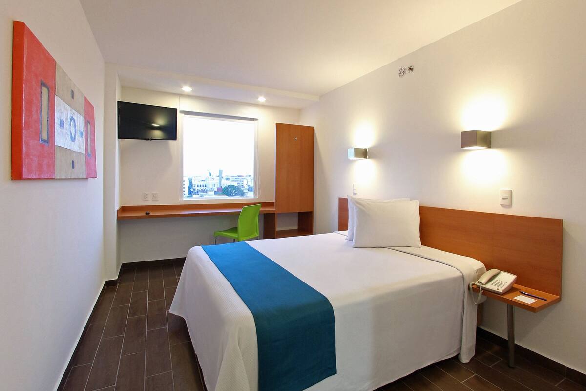 A hotel room with a bed, a desk, a chair, a TV, and a window with a city view.