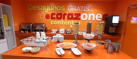 A buffet setup with various food items and utensils, a coffee machine, and a TV screen in the background.