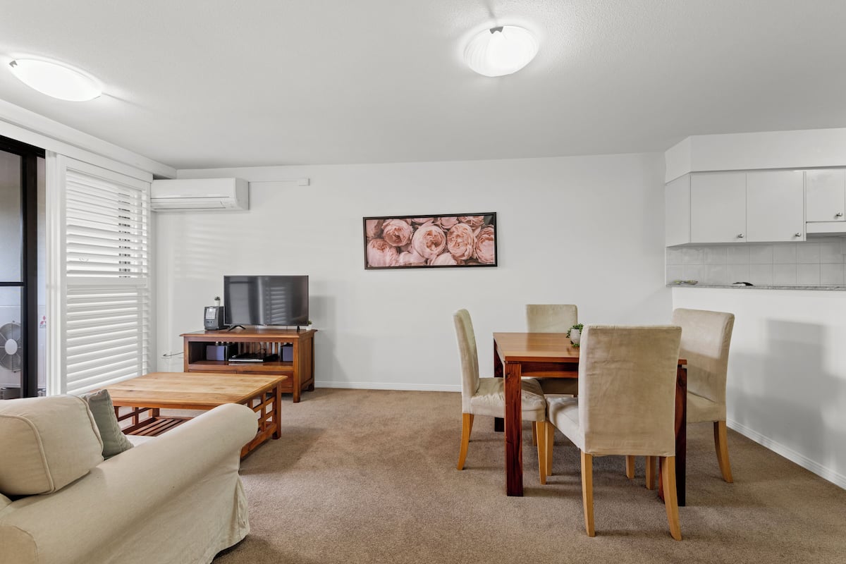 A modern living room with a sofa, a wooden coffee table, a dining table with chairs, a flat-screen TV, and a kitchen area with cabinets.