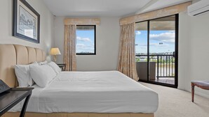Quarto de hotel com uma cama grande, uma escrivaninha com um laptop, uma luminária, um quadro na parede e uma varanda com vista para a cidade.
