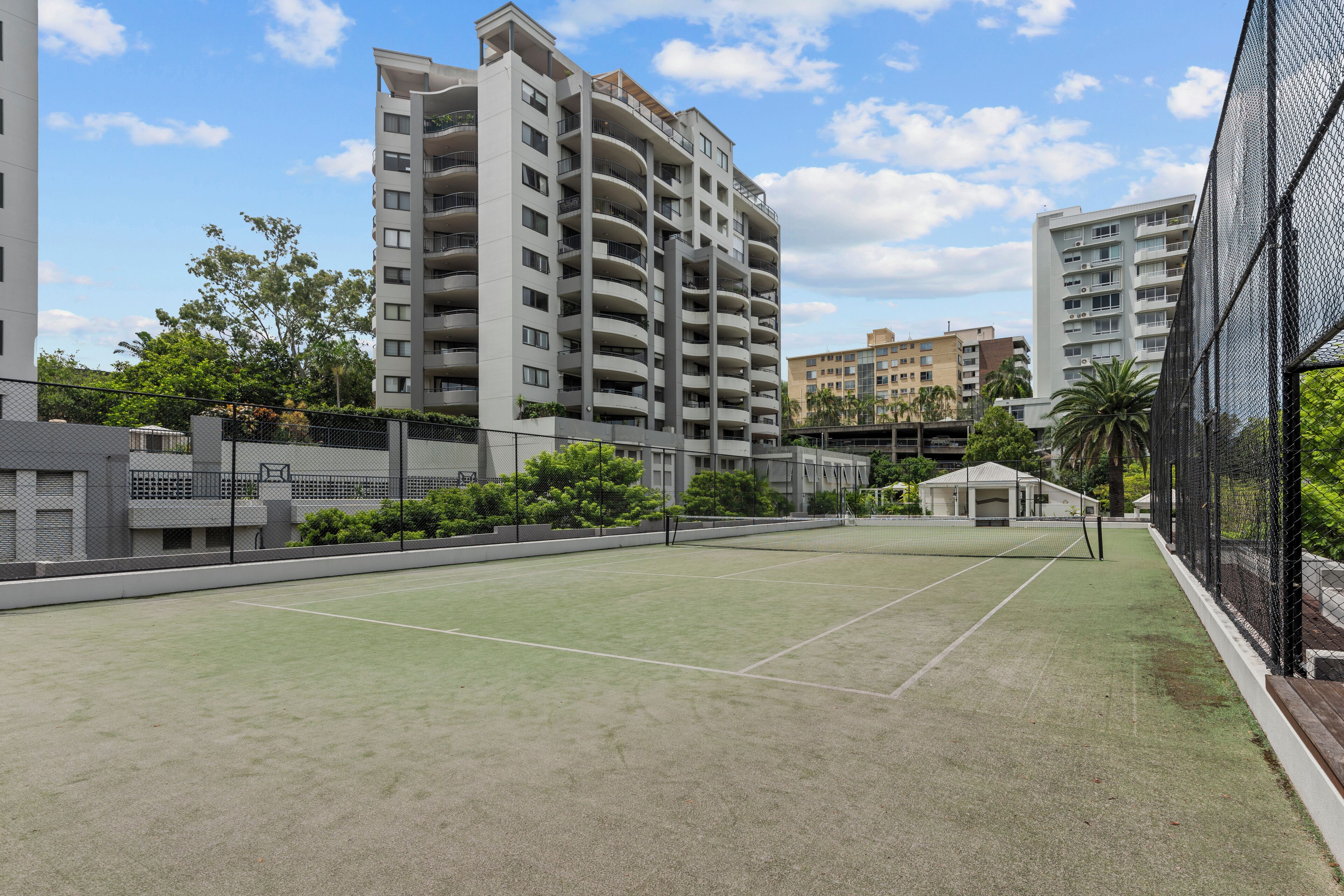 A tennis court surrounded by residential buildings, with a clear sky and some greenery.