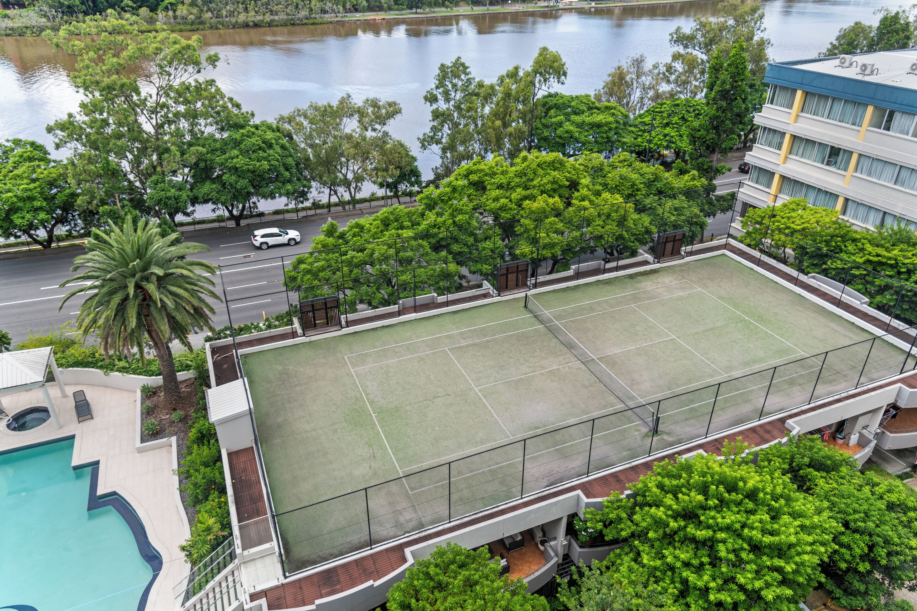 Una cancha de tenis con valla perimetral, una piscina y abundante vegetación.