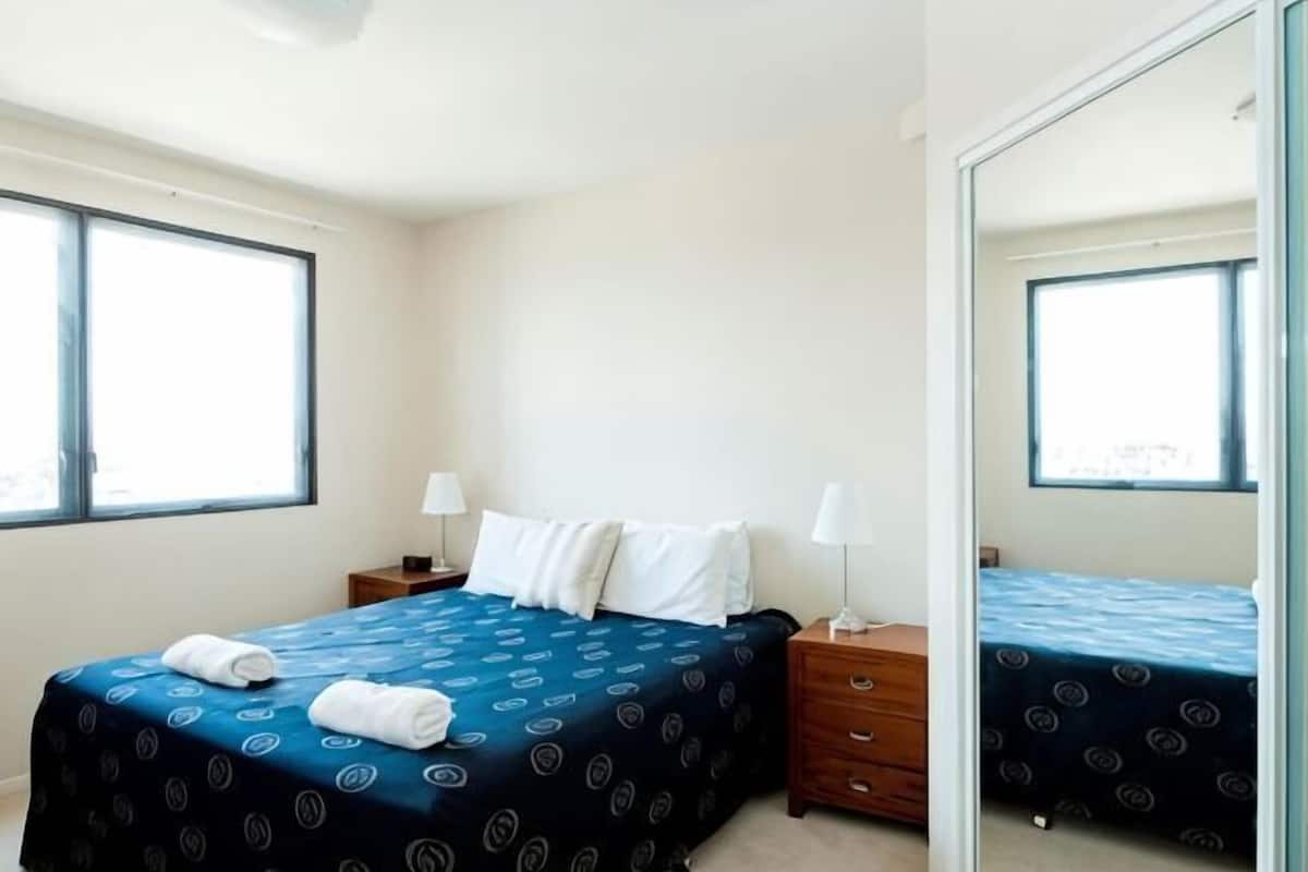 A hotel room with a bed, bedside tables, a mirror, and large windows.