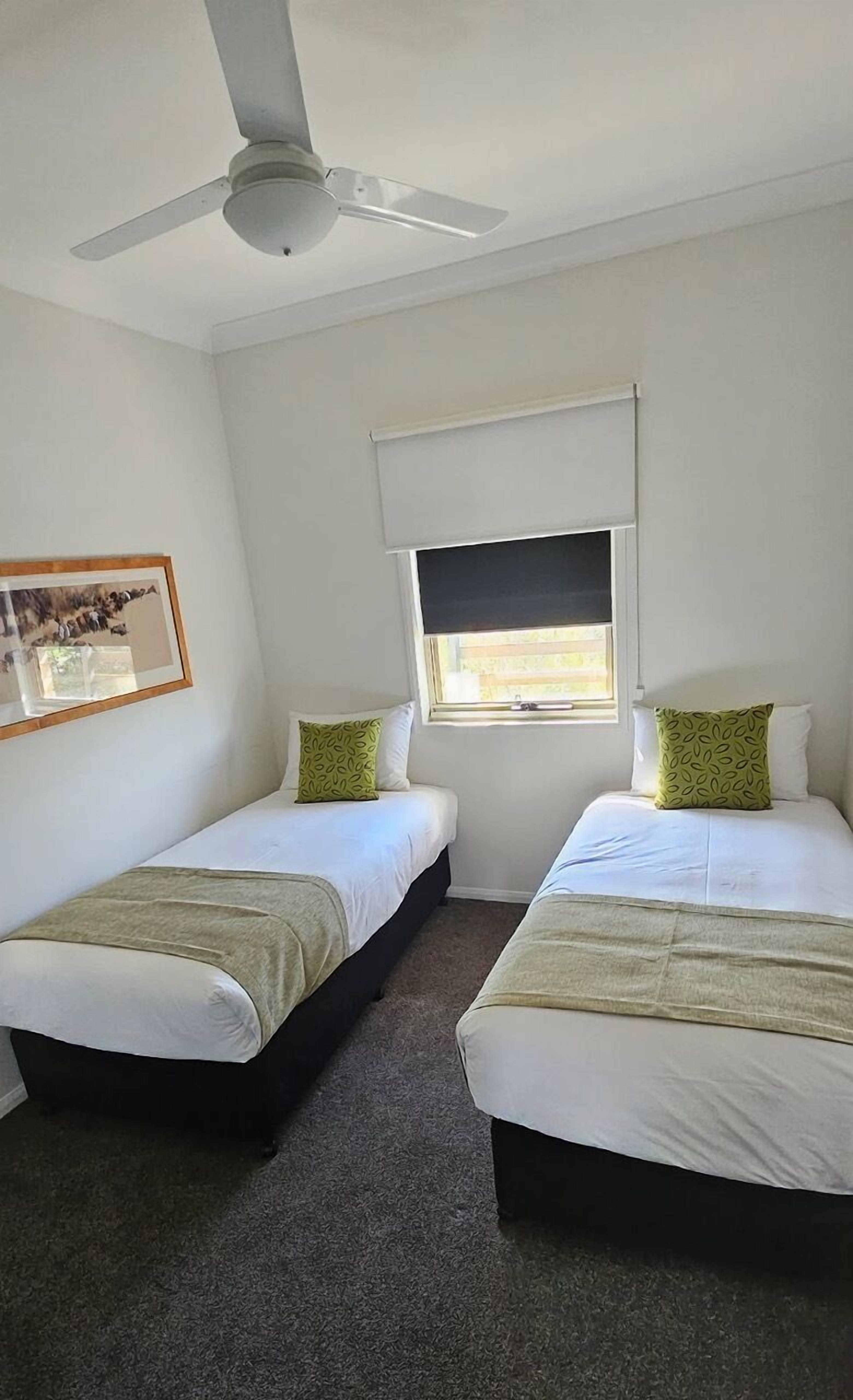 A room with two single beds, a ceiling fan, a window with a blind, and a framed picture on the wall.