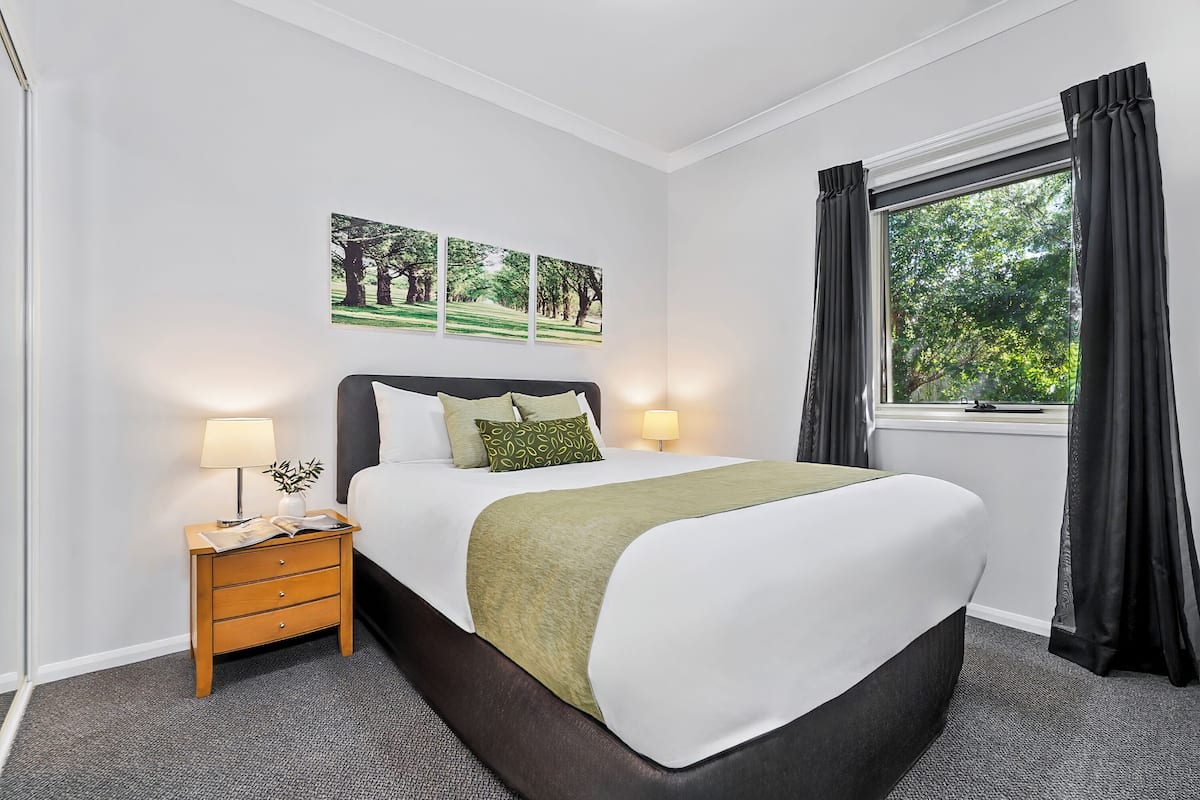 A neatly made bed with a green throw, two bedside lamps, a nightstand with a lamp, and a window with curtains.