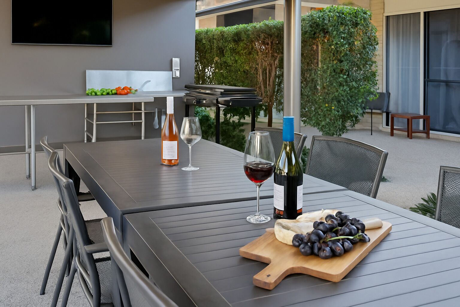 An outdoor dining area with a table set for two, featuring a bottle of wine, a glass, and a wooden cutting board with grapes.