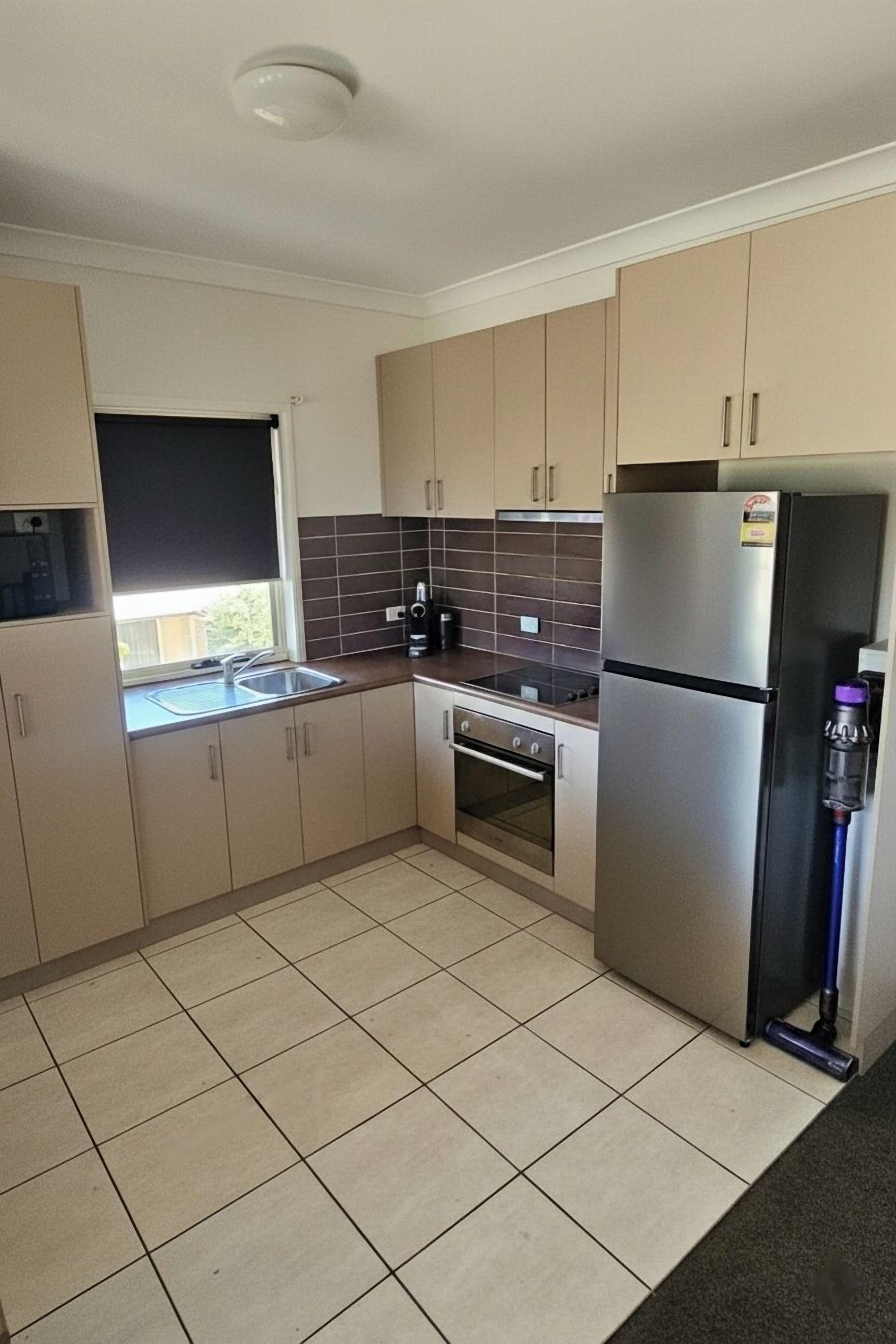 A modern kitchen with beige cabinets, a stainless steel refrigerator, a built-in oven, and a window with a black blind.