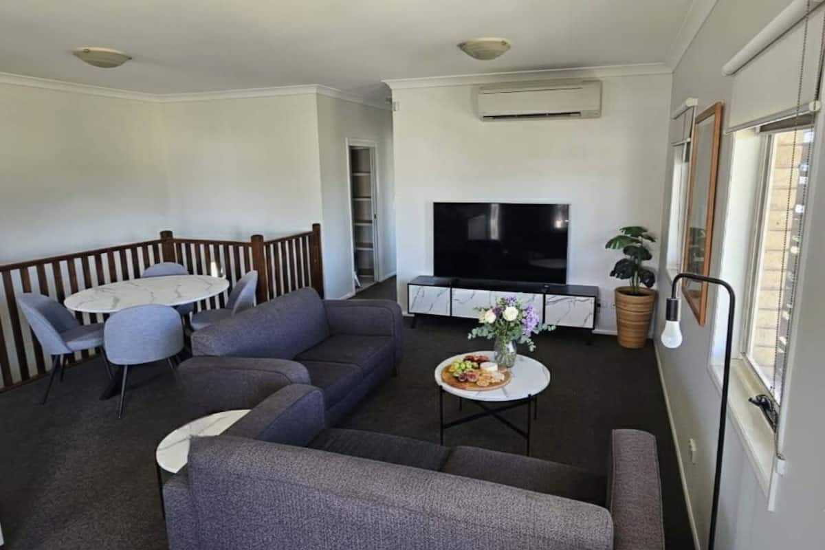 A living room with a gray sofa, a round table, a flat-screen TV, and a potted plant.