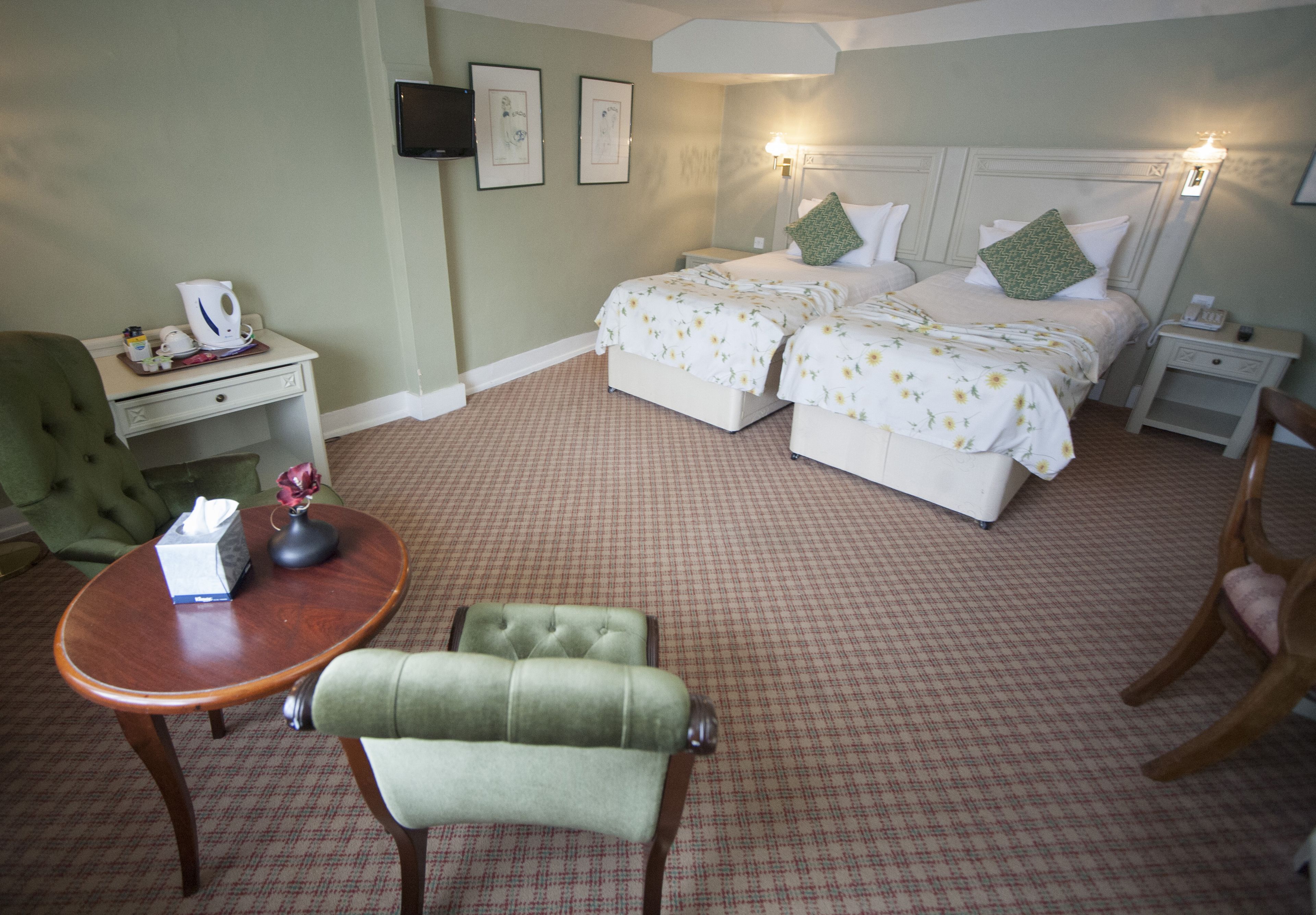 A hotel room with two beds, a green armchair, a wooden table, and a nightstand with a lamp.