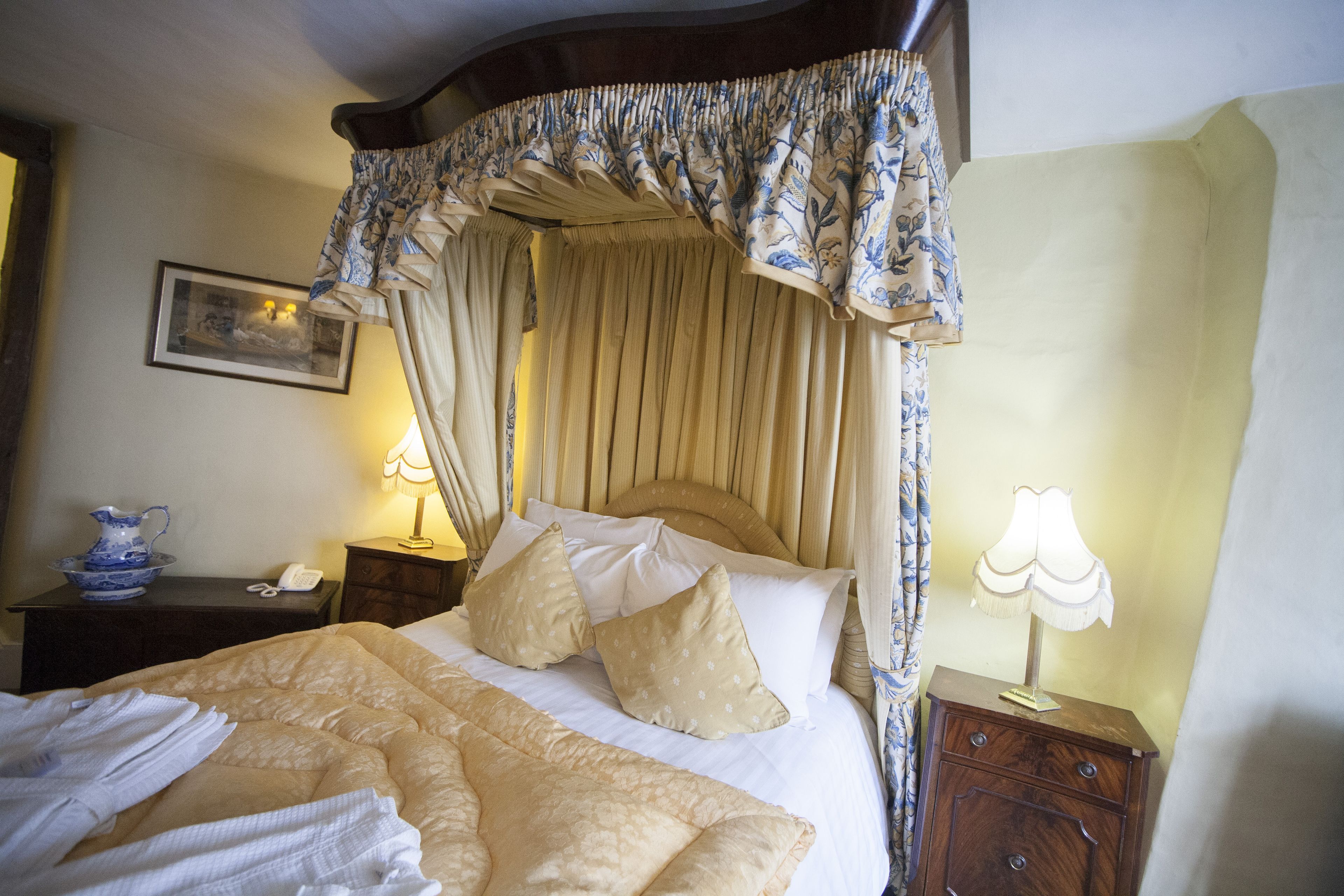 A neatly made bed with a canopy, a bedside table with a lamp, and a framed picture on the wall.