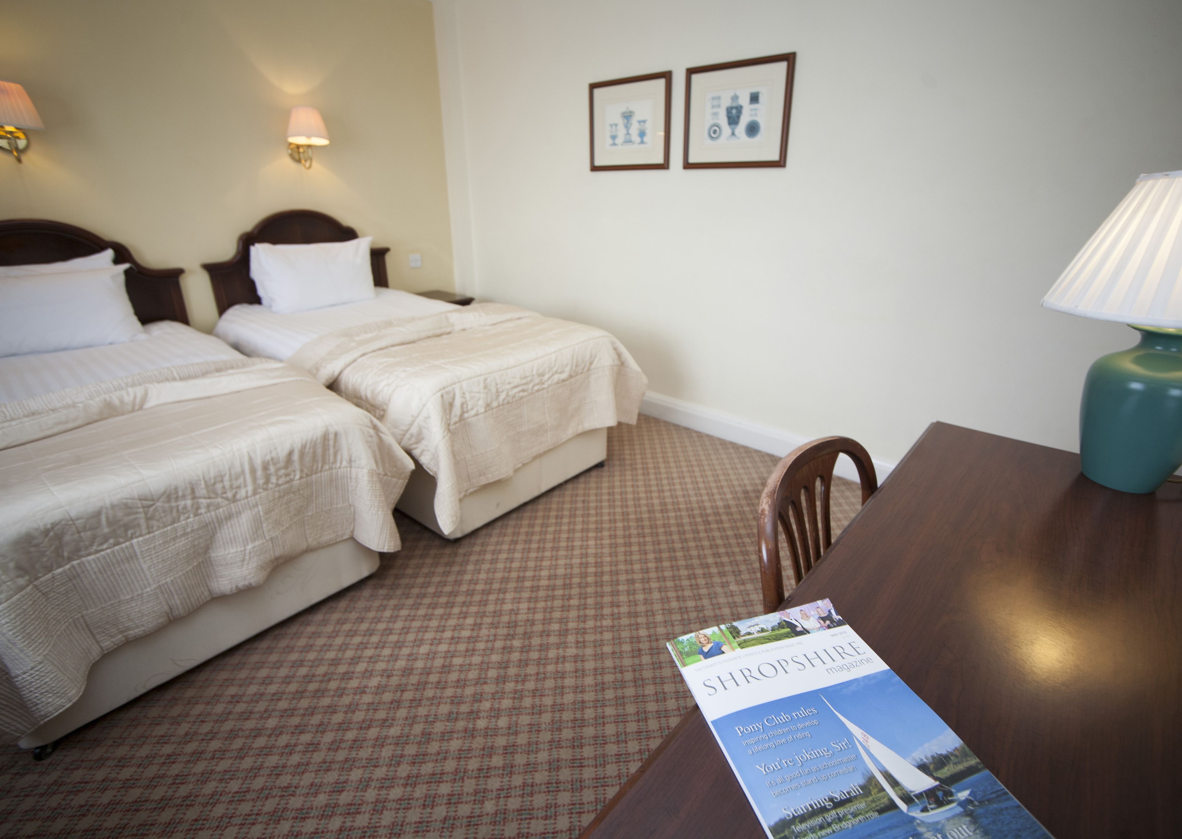A hotel room with two beds, a wooden table, a chair, a lamp, and a magazine on the table.