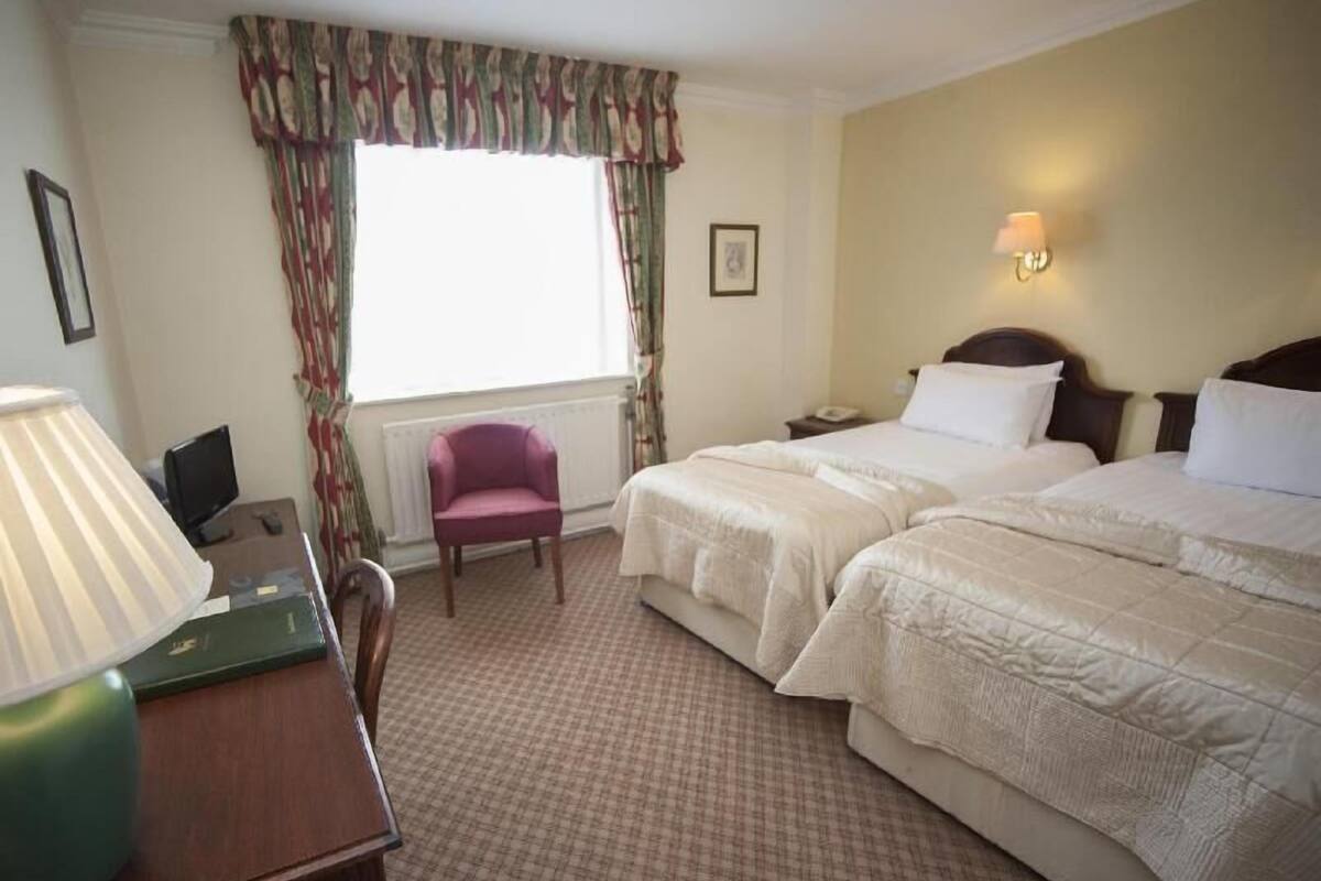 Habitación de hotel con dos camas, un escritorio con lámpara, una silla, un ventanal con cortinas y una luz en la pared.