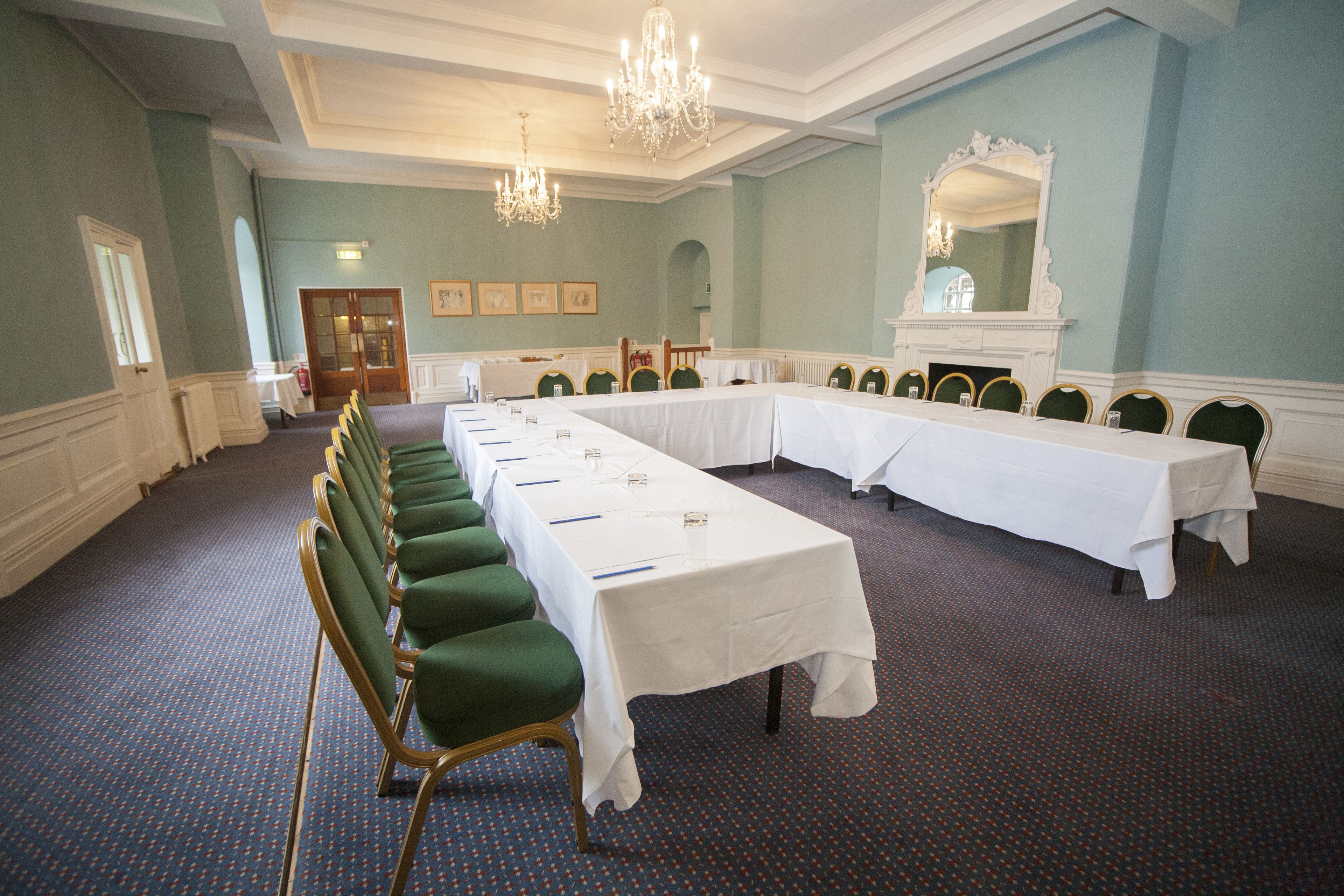 Un salón de conferencias con mesas largas, sillas verdes, candelabros y un espejo grande.
