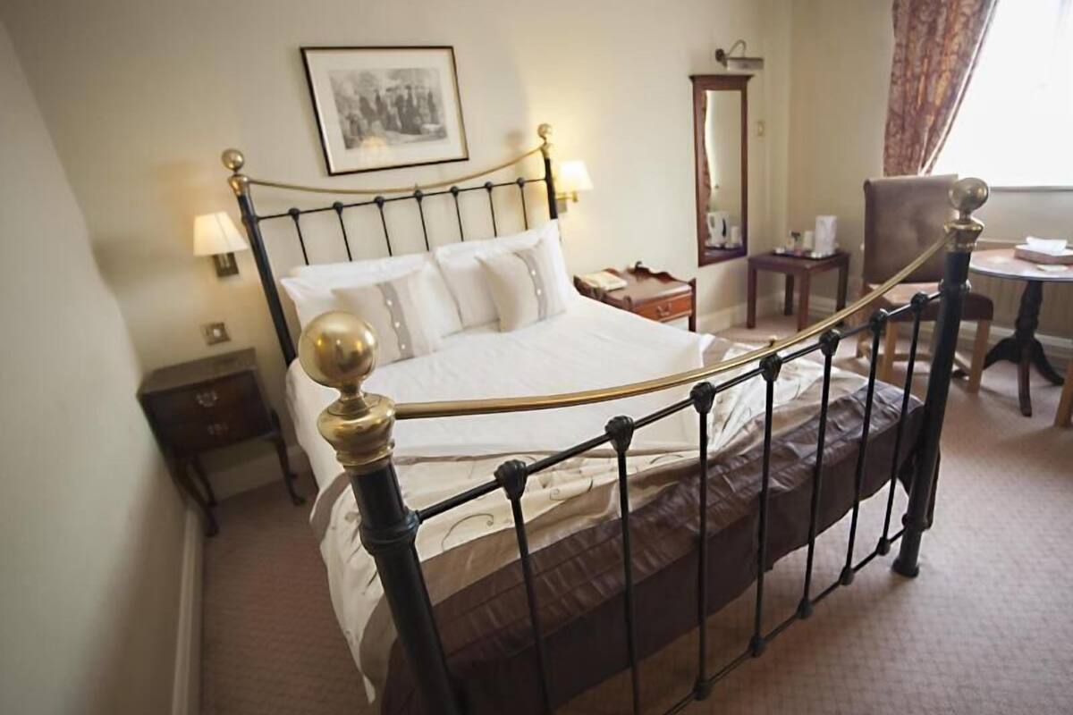 Una cama con estructura metálica y cabecera con un remate decorativo, una mesita de noche, una silla, un espejo y un cuadro enmarcado en la pared.
