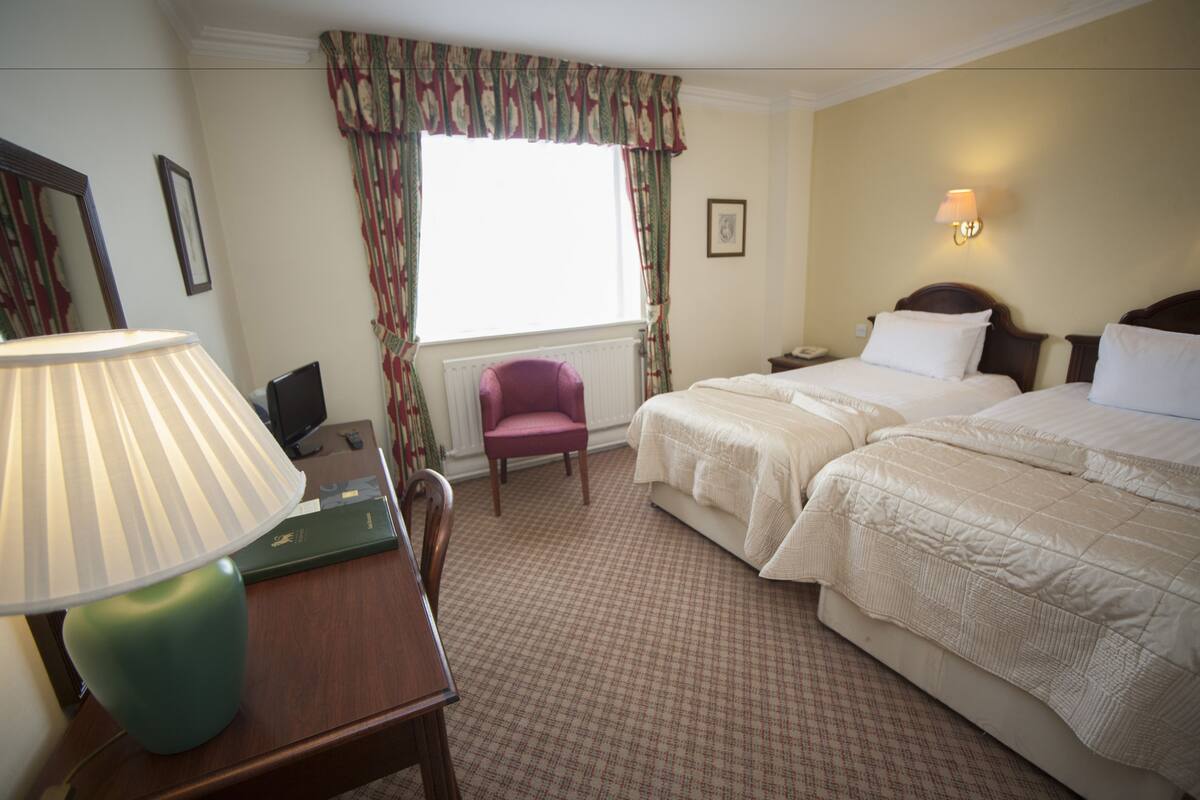 Habitación de hotel con dos camas, un escritorio con lámpara, una silla, una ventana con cortinas y una mesita de noche con teléfono.
