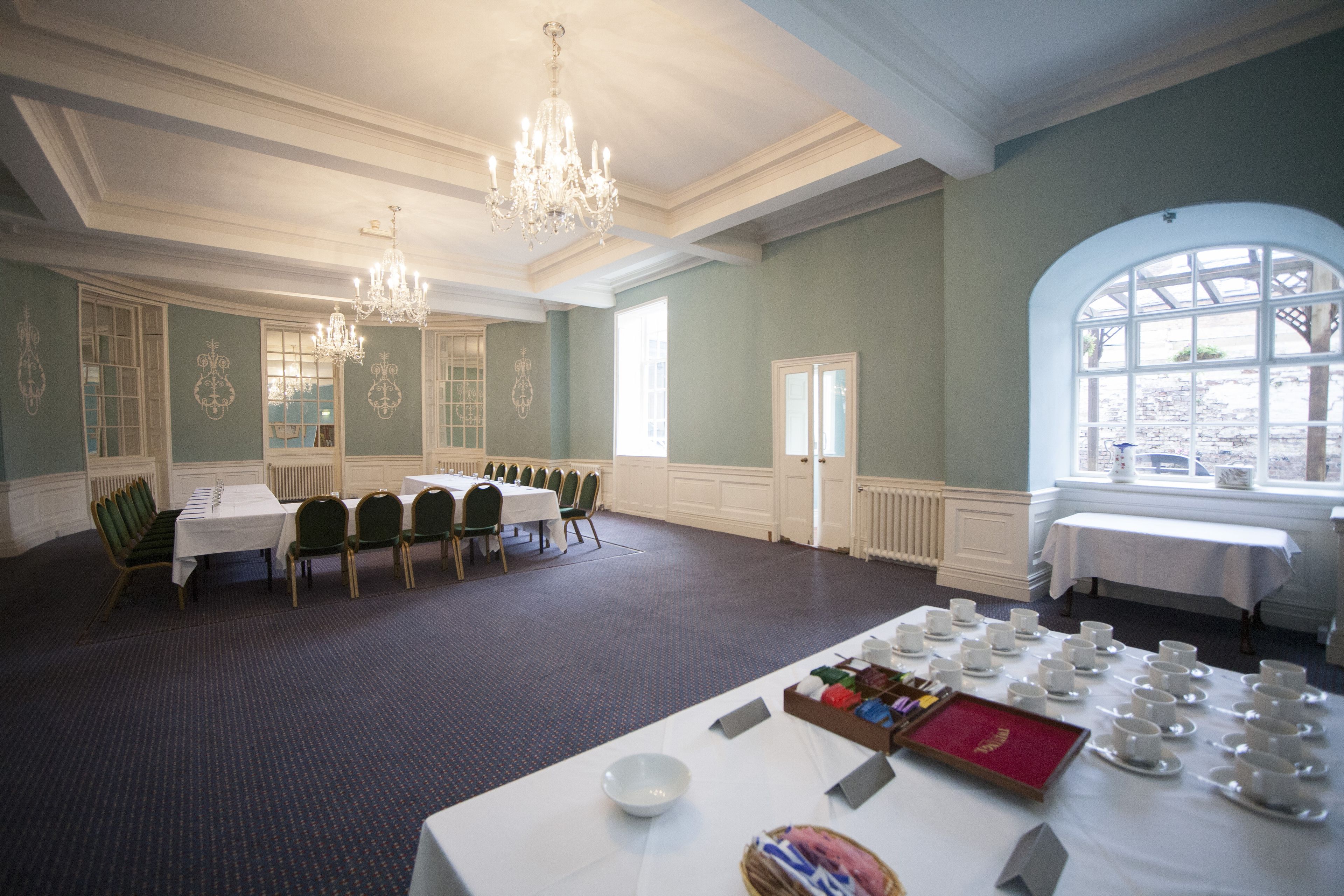 A formal dining room with tables set for a meal, chandeliers, and a large arched window.